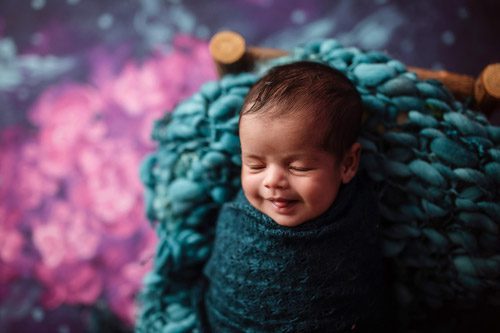 Joyful newborn wrapped in deep blue, lying on a plush teal knit against a vivid pink floral backdrop, photographed in Canberra