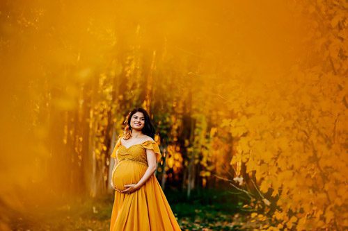 Expectant mother in a vibrant yellow dress stands amidst a stunning autumnal scene, photographed by a Canberra maternity photographer