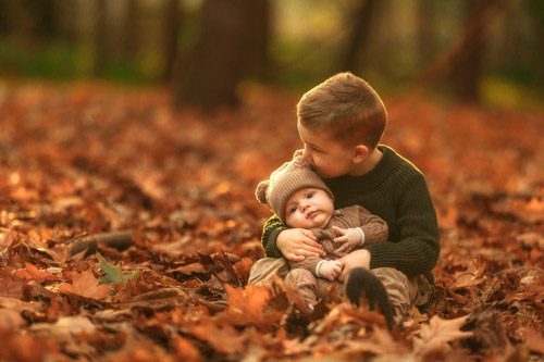 Older brother tenderly kissing his baby sibling amidst fallen leaves, captured by a winter Canberra family photographer