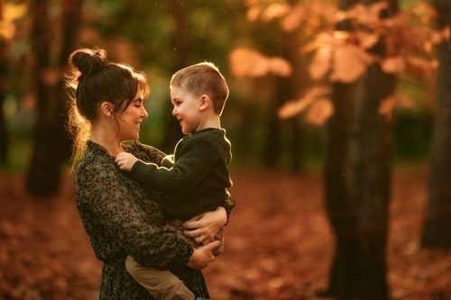 A mother holds her young son close, sharing a tender gaze amidst the rich, warm autumn leaves, captured by a winter Canberra family photographer.