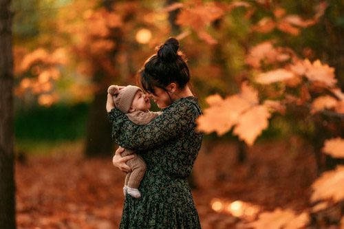 A mother lovingly lifts her baby in the air, surrounded by the golden hues of autumn, captured by a Canberra family photographer