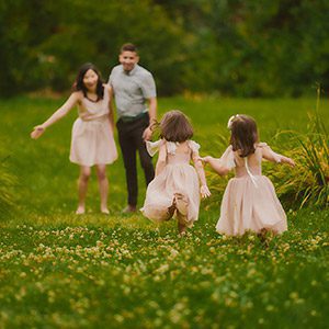 Spring-Summer-Family-Photos-Canberra_0002-copy Joyful family photo of two young girls running through a lush green field in Canberra, with parents smiling in the background, captured during a spring family session