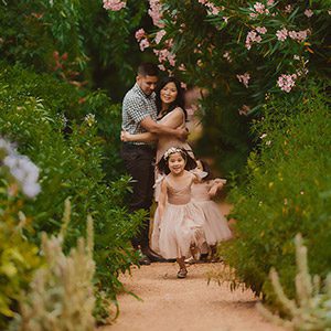 Beautiful summer family portrait in Canberra, with two young girls running along a garden path, surrounded by vibrant flowers, as their parents share a loving embrace